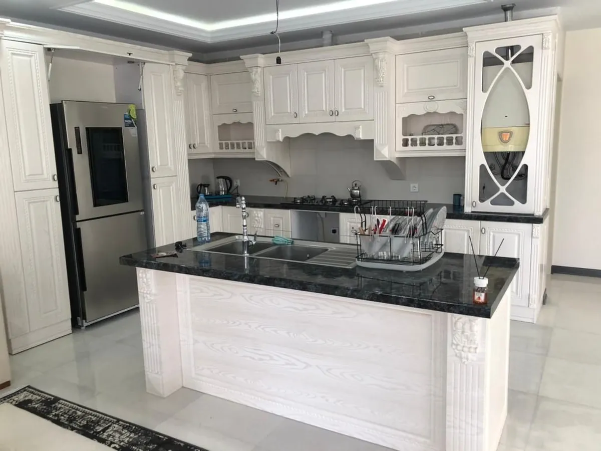 کابینت روکش چوب سفید صدفی با جزیره و دو کابینت کمدی در دو طرف باکس یخچال و کمد ویترینی در سمت راست